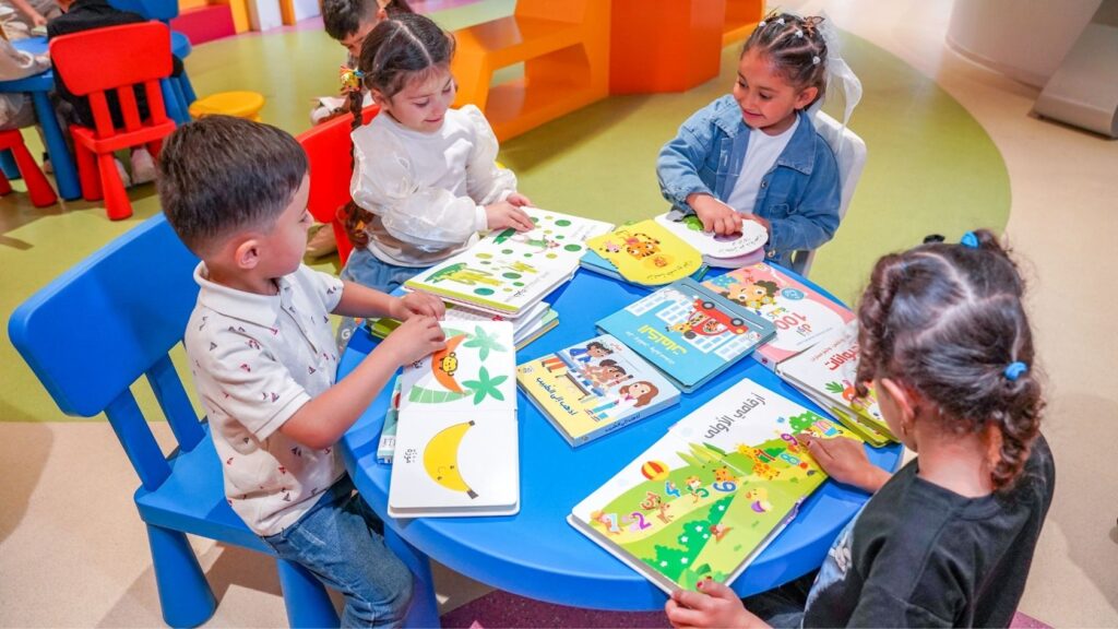 Quatre jeunes enfants lisent des livres illustrés autour d’une table dans un centre éducatif coloré
